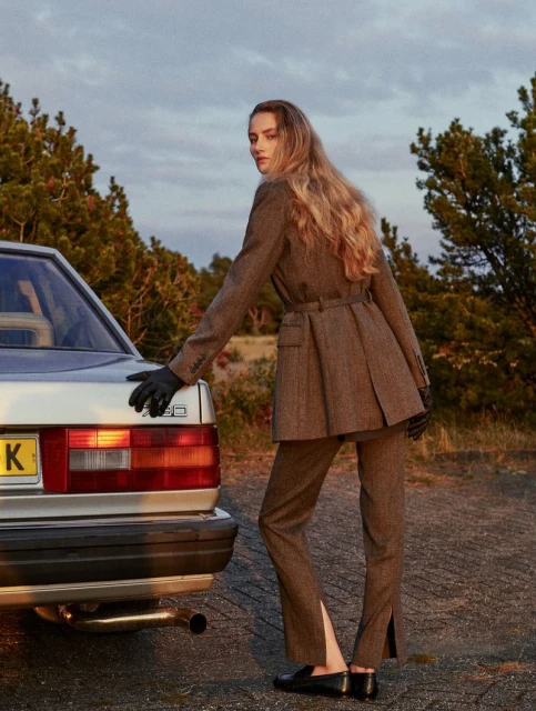 Woman in a brown suit and black gloves poses next to a silver car in an outdoor setting with green trees and a cloudy sky.