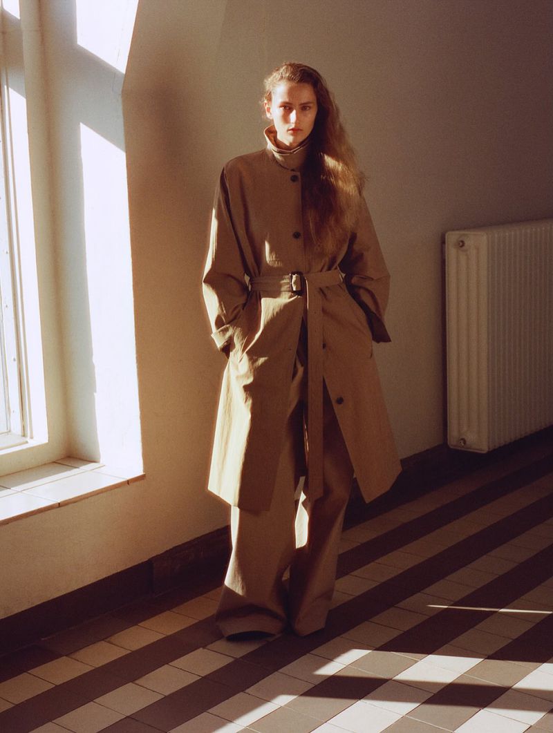 Woman in beige trench coat and wide-leg pants poses indoors near a window with shadows on floor; minimalist fashion editorial.