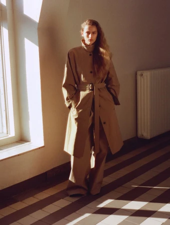 Woman in beige trench coat and wide-leg pants poses indoors near a window with shadows on floor; minimalist fashion editorial.