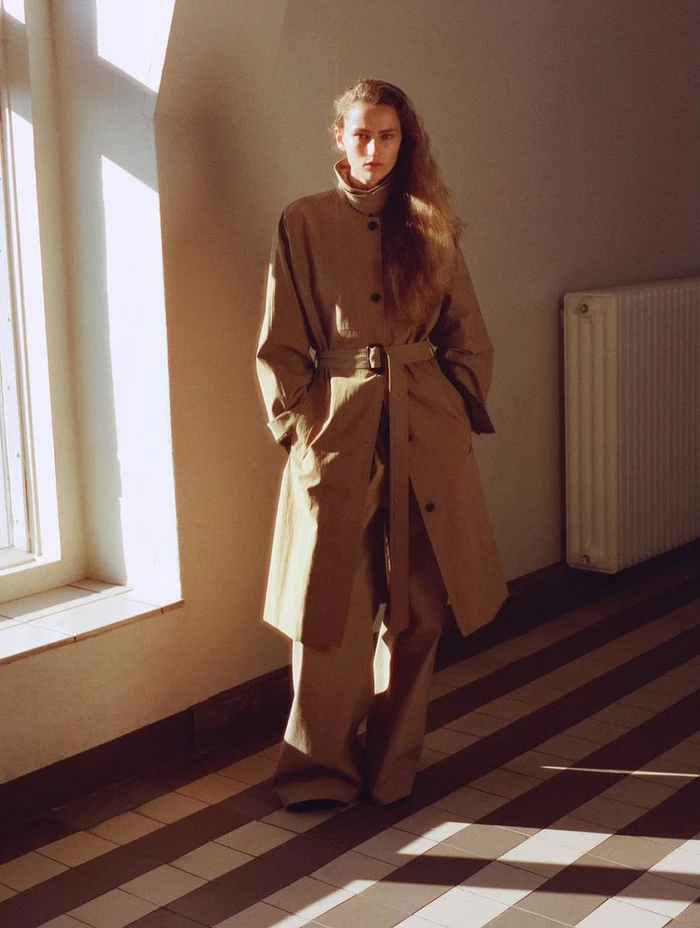 Model standing in a belted beige trench coat and wide trousers in a sunlit hallway with striped tile flooring.