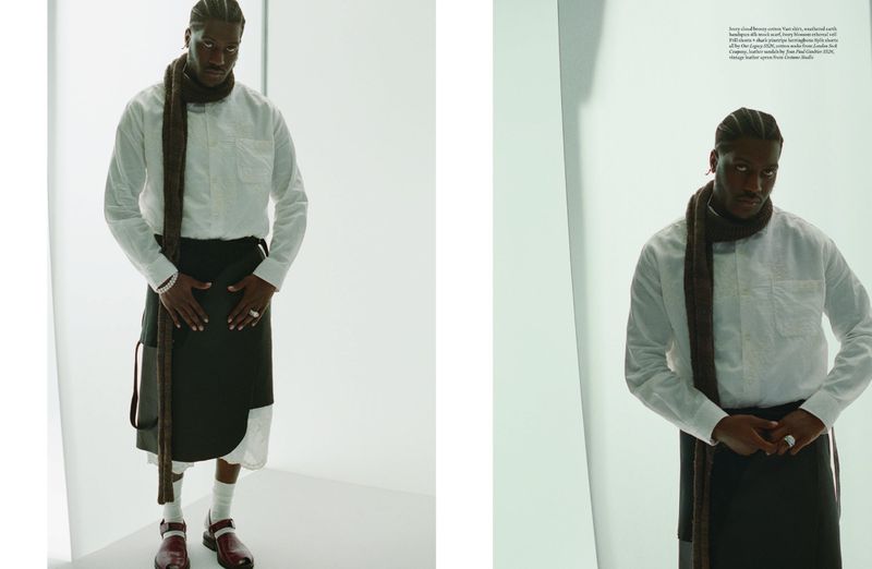An adult male model stands in a white photo studio wearing a white shirt, brown scarf, dark wrap skirt, white socks and maroon clogs.