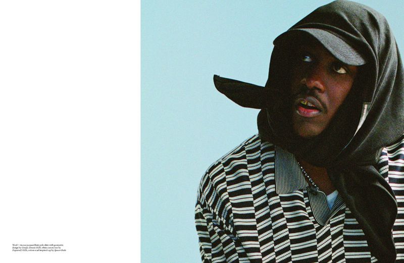 Portrait of a Black adult male wearing a striped polo shirt and dark hooded cap, looking to the side against a light blue background.