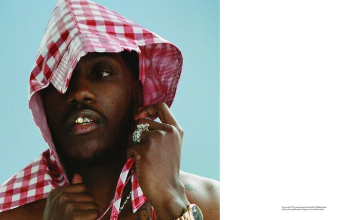 Lil Yachty wearing a red and white gingham hood, gold grillz, and a diamond ring against a solid pale blue background.