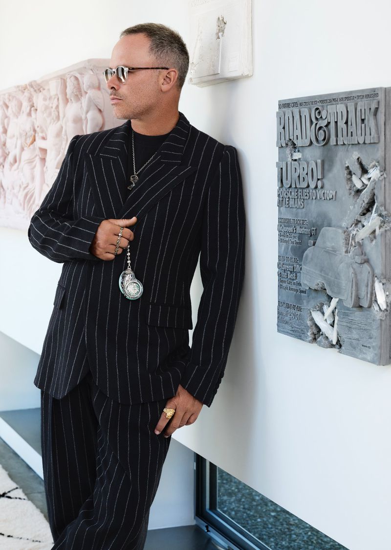 Stylish man in pinstripe suit poses indoors near concrete art. He wears sunglasses, jewelry, and holds a fob watch, embodying fashion and style.