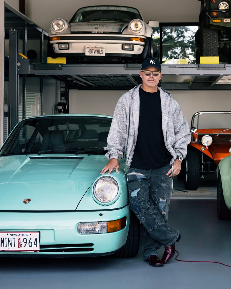 Adult male leans on a light blue Porsche in a garage with other vintage cars and vehicle lift in the background.