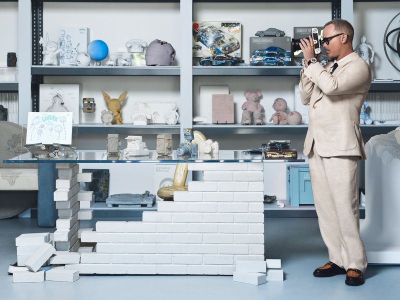 Adult male in tan suit stands in front of shelves with collectibles while holding a camera, set against a white backdrop.