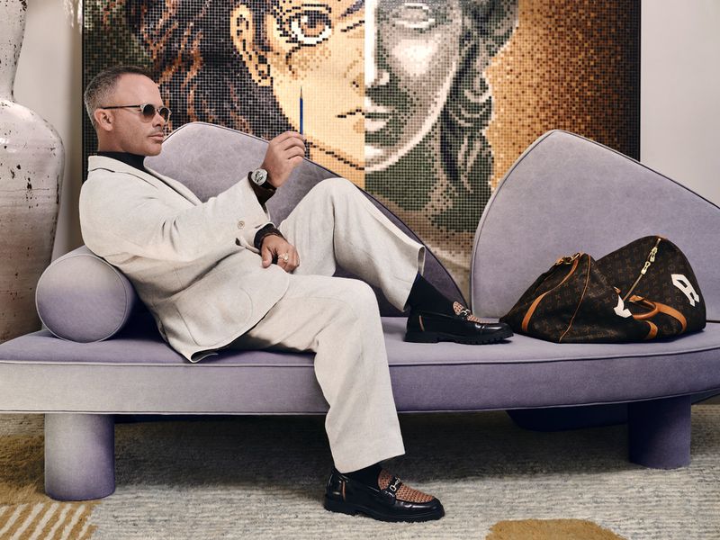 Stylish adult male seated indoors on modern lilac couch, wearing sunglasses and suit, next to Louis Vuitton bag, in front of mosaic art.
