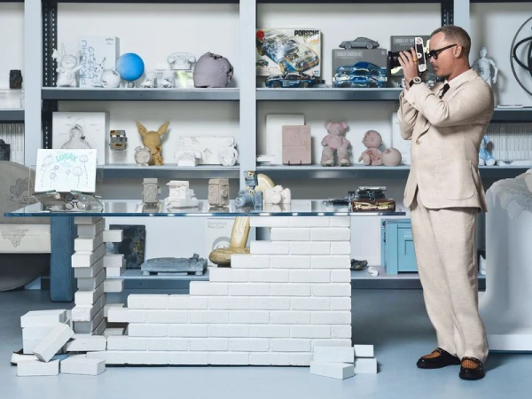 Artist Daniel Arsham in a beige linen suit holding a camera beside a white brick and glass desk in his art studio.