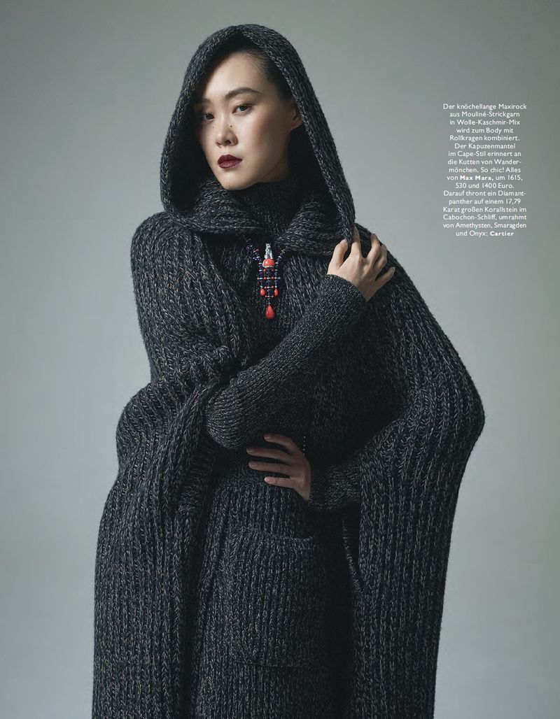 Portrait of an Asian woman wearing a grey hooded knit cape with a decorative pin, set against a light grey studio backdrop.