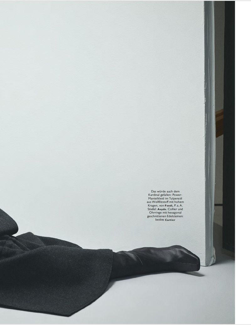 Fashion editorial shot of a dark gray coat and glove next to a white wall with text on it in a studio setting. 