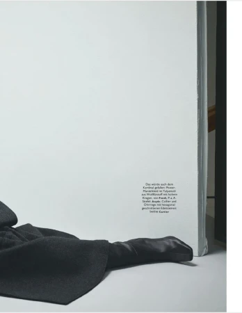 Fashion editorial shot of a dark gray coat and glove next to a white wall with text on it in a studio setting. 