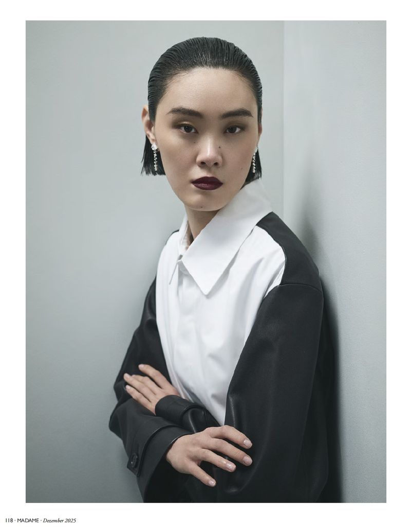 Elegant Asian woman in fashion shot with dark hair, wearing a white shirt with black sleeves, posing indoors against a gray wall.