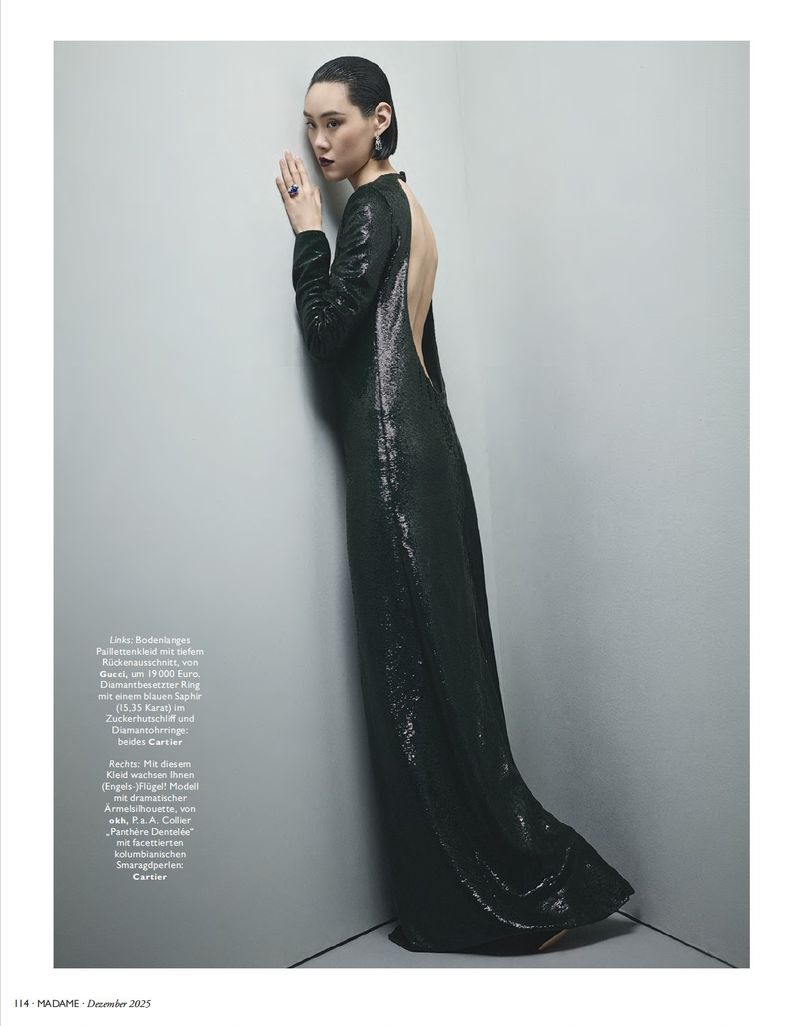 Elegant adult female fashion model poses in a full-length gown with a backless design and long sleeves in a minimalist studio corner setting.