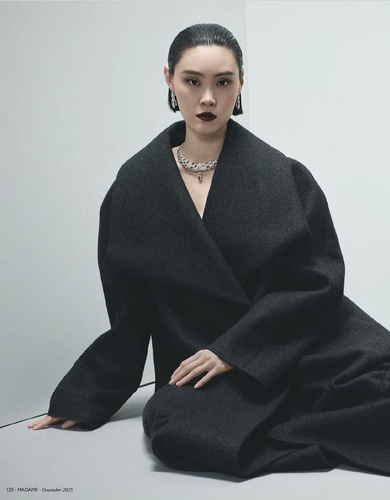 An Asian woman is elegantly posed in a dark overcoat with jewelry against a minimalist background, showcasing high fashion and beauty.