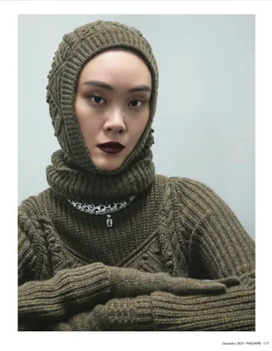 Model wearing an olive green knitted balaclava and sweater with a patterned necklace against a plain grey background