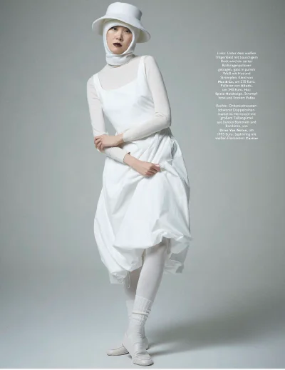 Model wearing an all-white outfit including a hat, turtleneck, and voluminous dress against a neutral grey studio background.