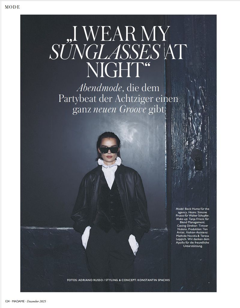 A woman poses in a black suit and sunglasses, styled for a fashion editorial with a backdrop featuring a text overlay and dark door.
