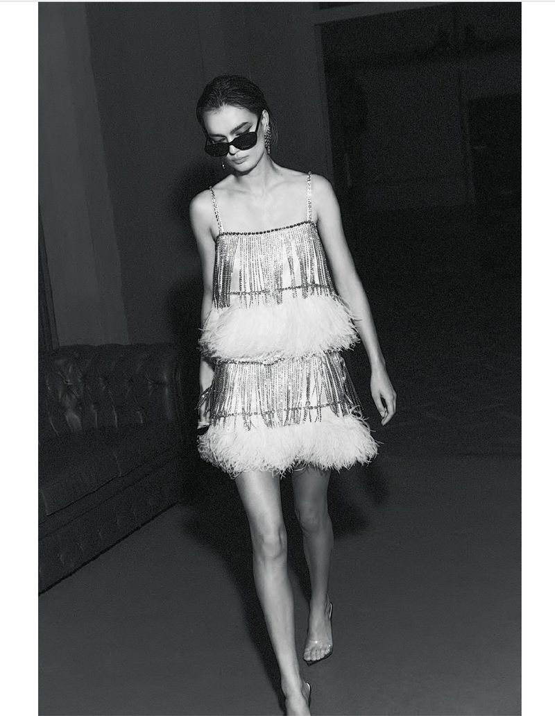 A woman wearing a silver fringe dress with white feathers and sunglasses walks in a dimly lit interior space with a couch.
