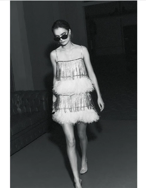 A woman wearing a silver fringe dress with white feathers and sunglasses walks in a dimly lit interior space with a couch.