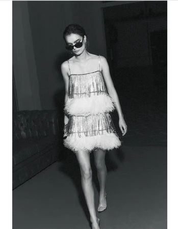A woman wearing a silver fringe dress with white feathers and sunglasses walks in a dimly lit interior space with a couch.