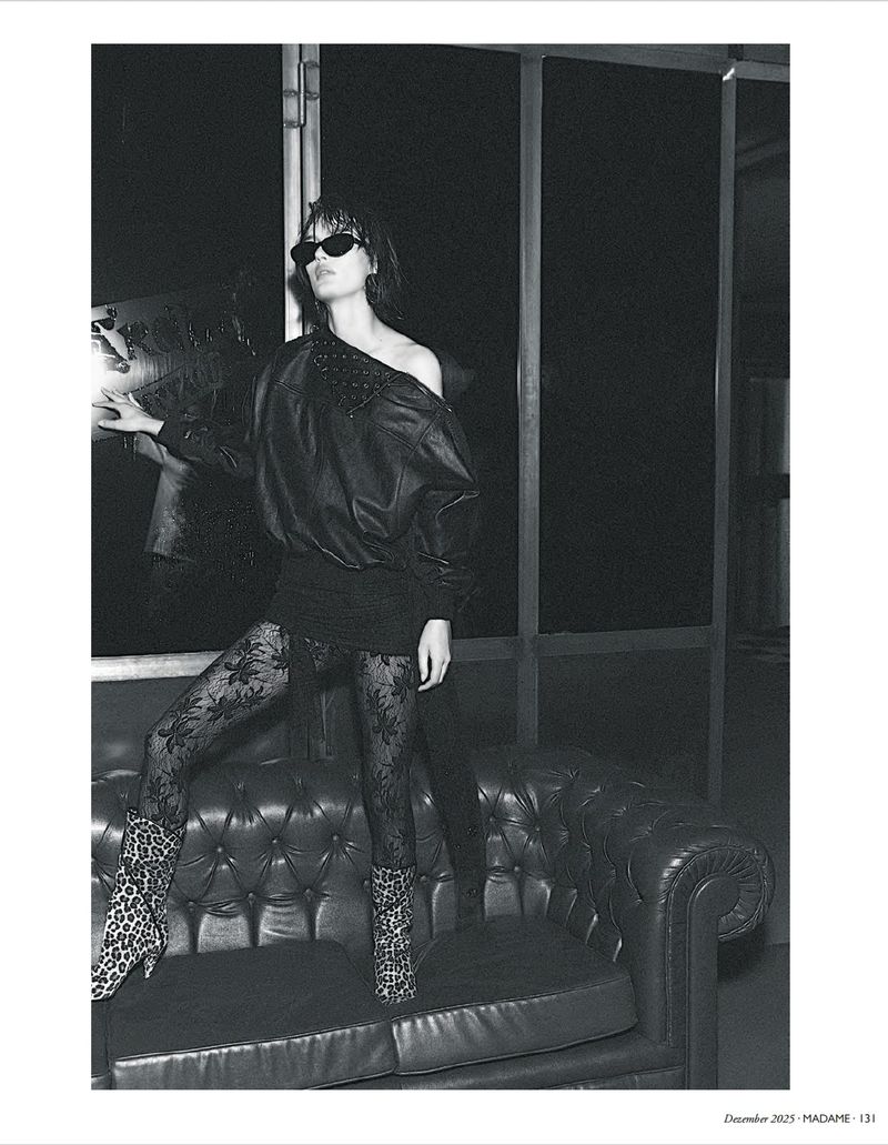 Black-and-white fashion shot of a woman in sunglasses, lace tights, and animal-print boots, posing on a black leather sofa near a window.