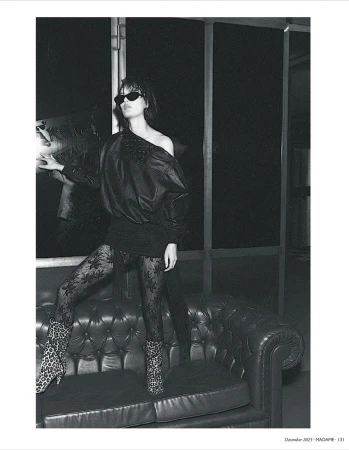 Black-and-white fashion shot of a woman in sunglasses, lace tights, and animal-print boots, posing on a black leather sofa near a window.