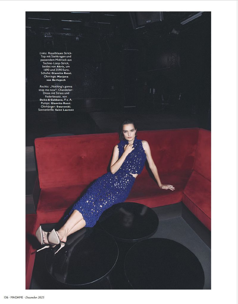 A woman in a blue sequined two-piece dress sits on a red couch in an indoor fashion setting, with black round tables nearby.