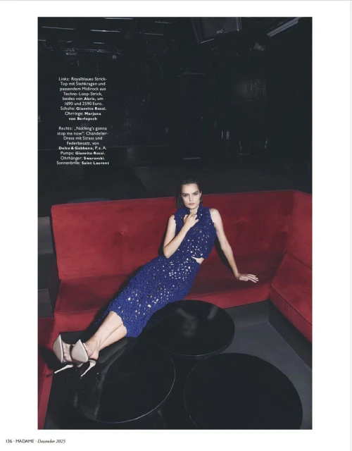 A woman in a blue sequined two-piece dress sits on a red couch in an indoor fashion setting, with black round tables nearby.