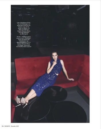 A woman in a blue sequined two-piece dress sits on a red couch in an indoor fashion setting, with black round tables nearby.