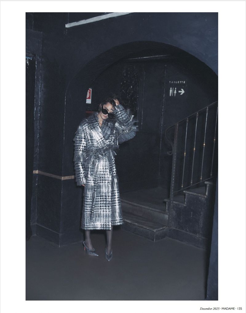 Woman in metallic plaid coat posing in dark archway near stairs. Fashion shot featuring sunglasses, heels, and cool toned aesthetic.