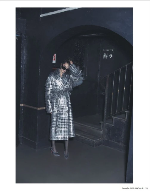 Woman in metallic plaid coat posing in dark archway near stairs. Fashion shot featuring sunglasses, heels, and cool toned aesthetic.