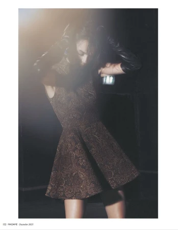 Woman poses in a patterned sleeveless dress with leather sleeves against a dark background with a bright light flare in a fashion shoot.