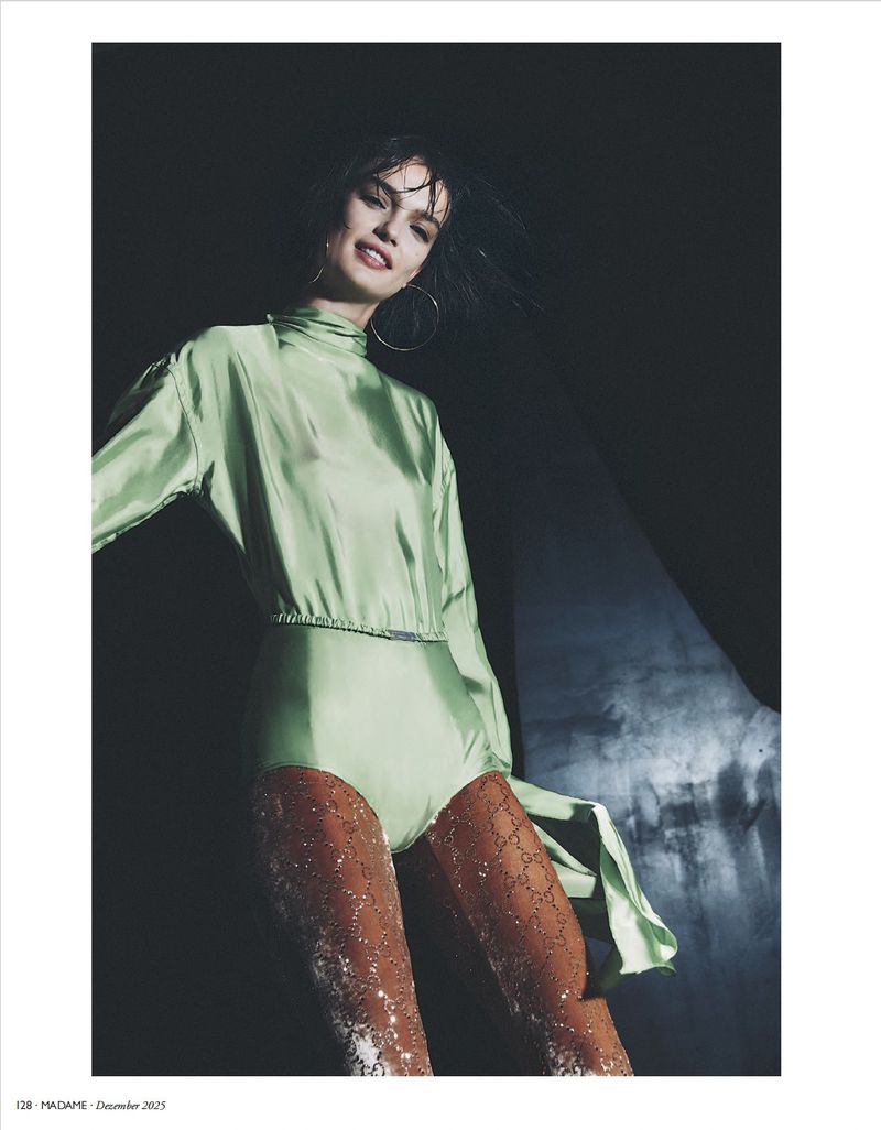 Fashion editorial shot of a woman in a pale green, shiny bodysuit with sparkling tights, posing in front of a dark background.