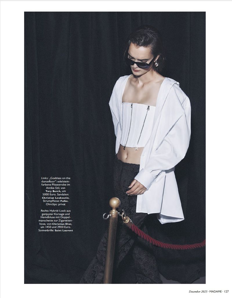 Fashion editorial shot of a woman with sunglasses in a white open shirt, corset, and black patterned pants in a dark studio.
