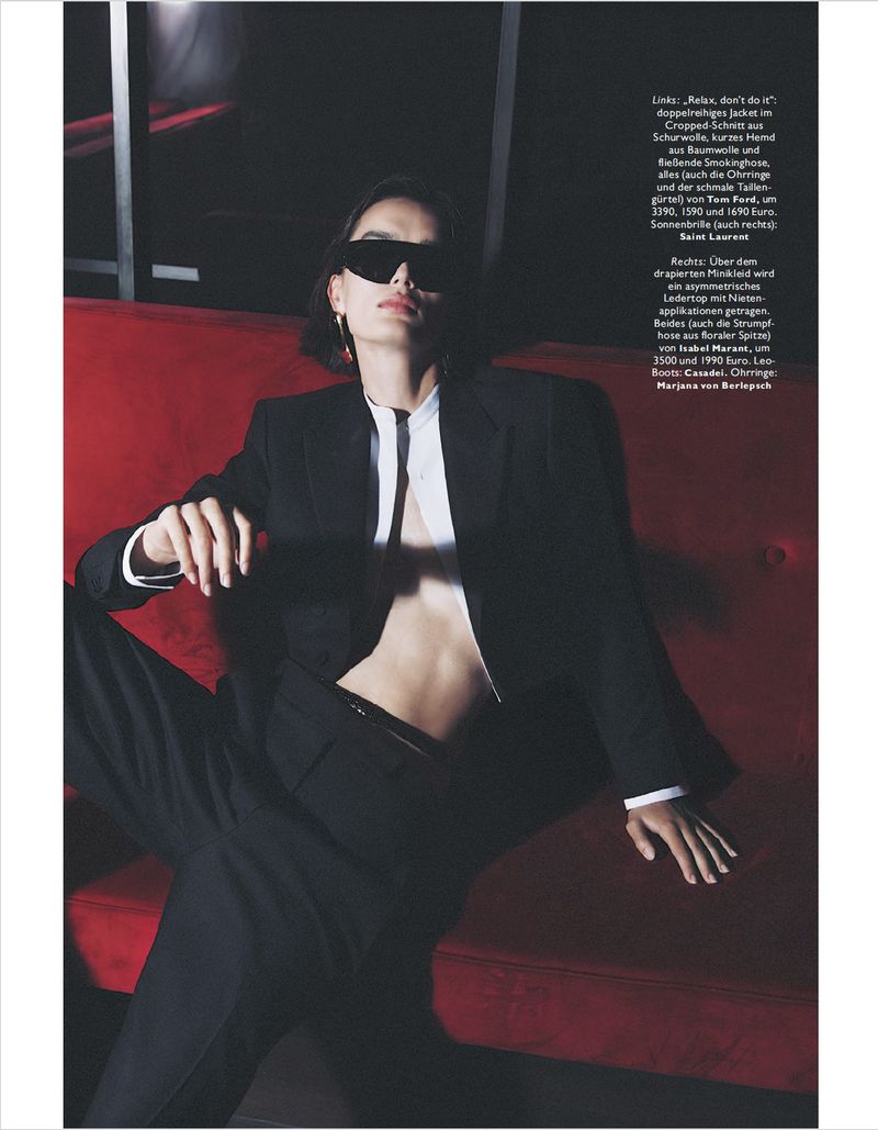 Stylish woman in sunglasses and a black suit poses on a red couch against a dark background in a fashion editorial shot.