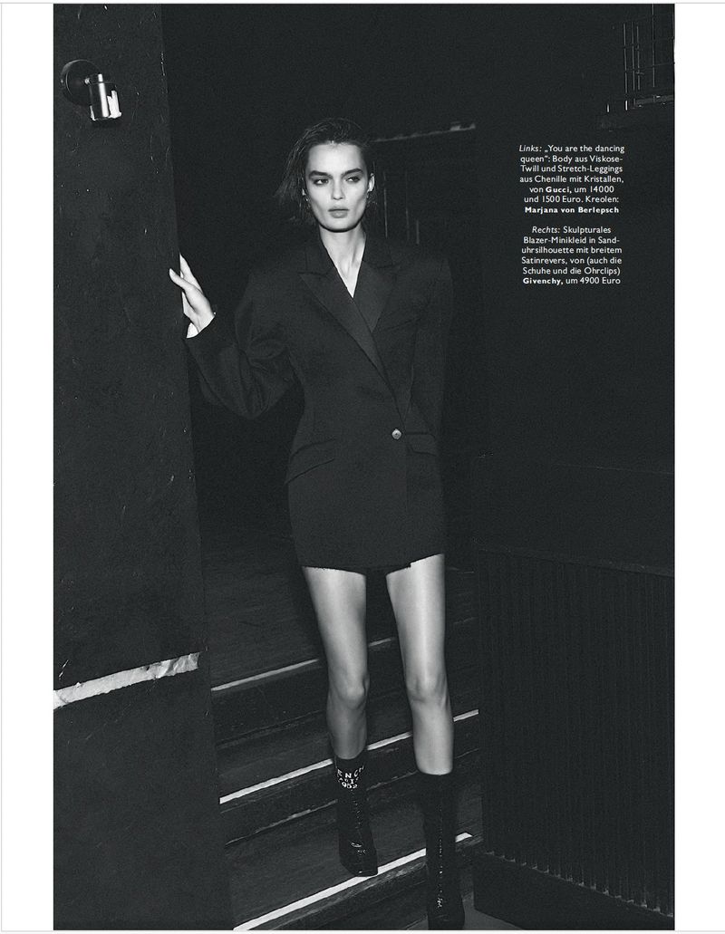 A woman in a black jacket dress and boots poses on dark steps, creating a fashion-forward editorial image with monochrome tones and modern style.