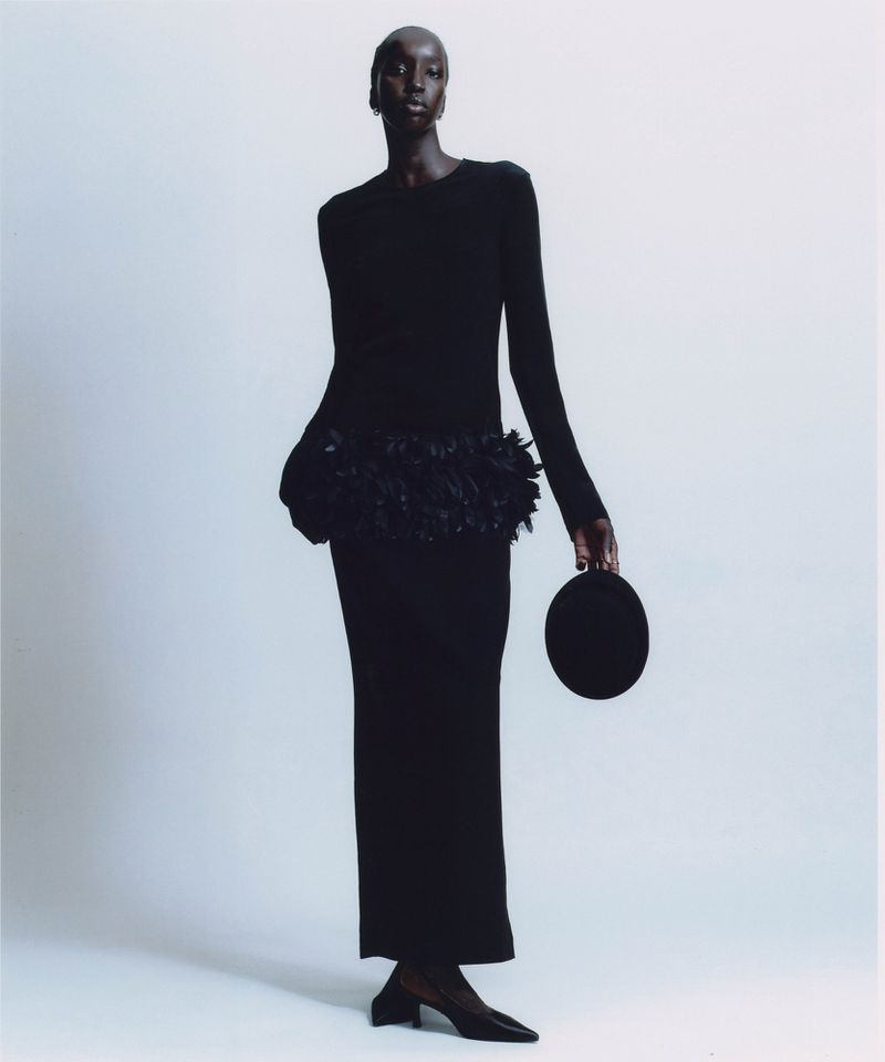 Full-length studio shot of a Black female fashion model wearing a long-sleeved black dress with feathered accent, holding a round purse.