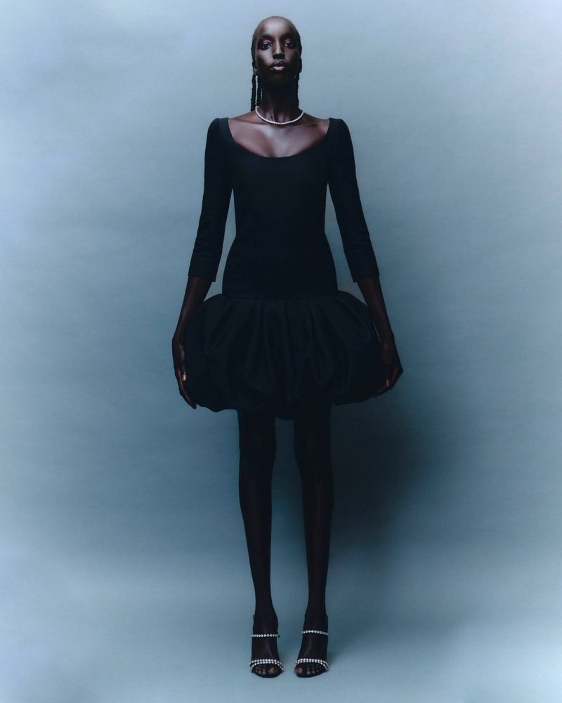 Full-length shot of a Black female model in a black dress with a voluminous skirt, posing in front of a light blue backdrop.