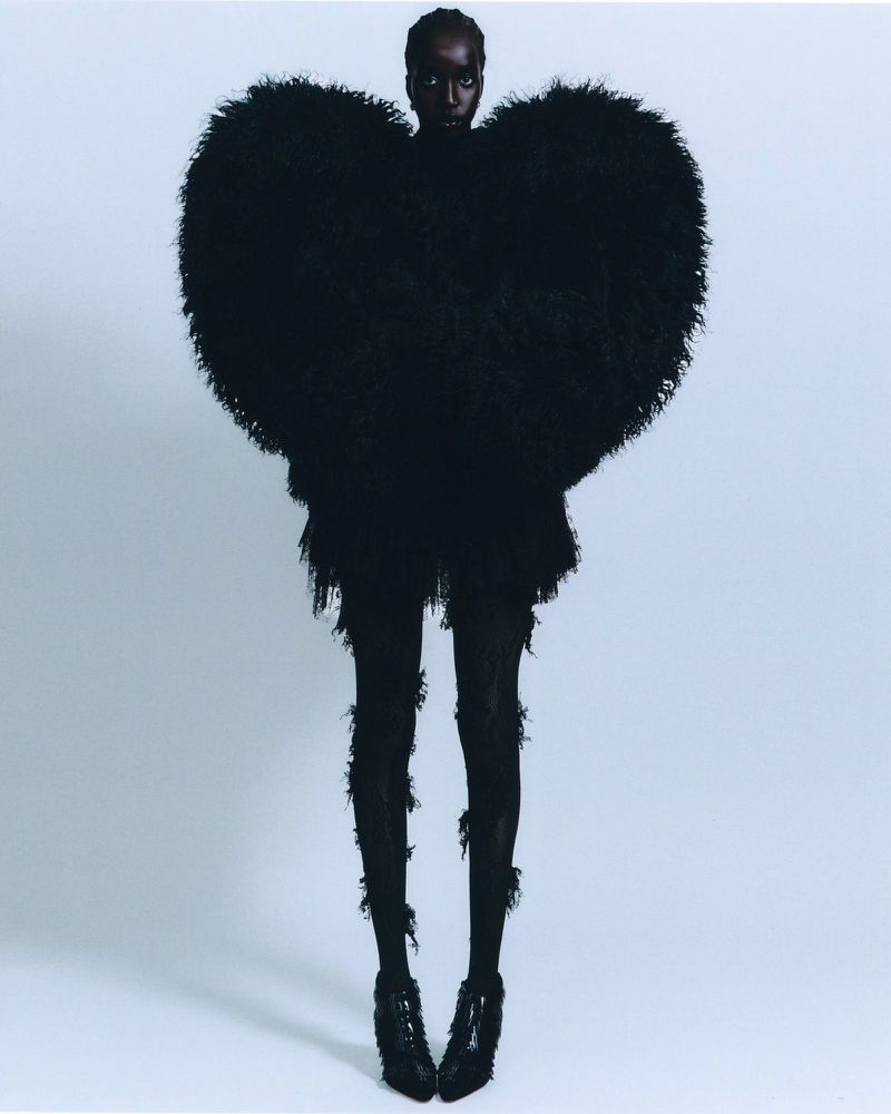 Fashionable woman poses in an oversized black heart-shaped dress with matching tights and heels against a pale blue gradient background.