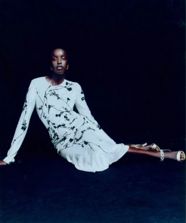Elegant Black woman in a white floral dress sits against a dark backdrop, accessorized with gold shoes and bright makeup, posing for a fashion shot.