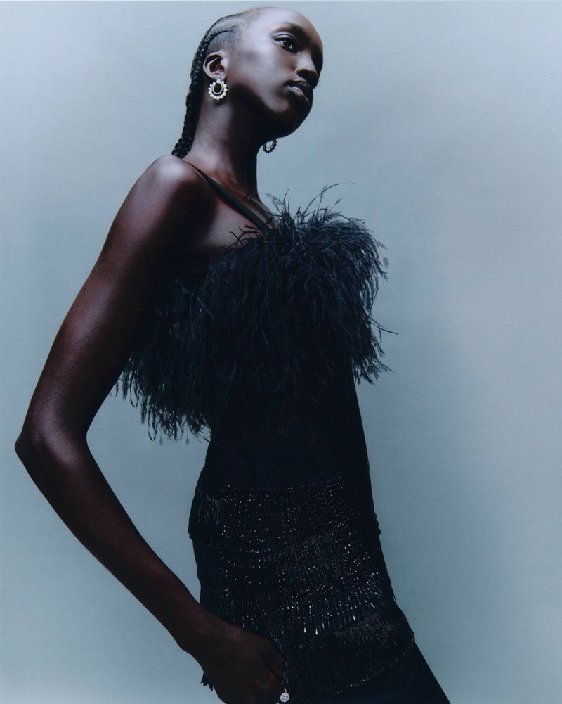 Headshot of an adult Black woman with braided hair wearing a black feather top and dress with sequin accents against a light blue background.
