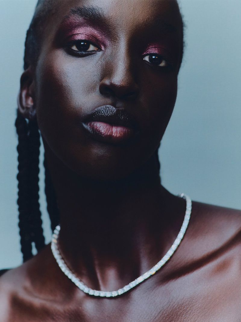 Close-up portrait of a dark-skinned adult woman with magenta eyeshadow and a white beaded necklace against a grey background.