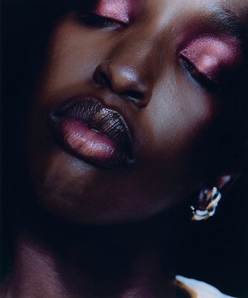 Close-up portrait of a Black woman with closed eyes and glittery pink makeup on the eyelids. She is wearing earrings, with dark lighting.