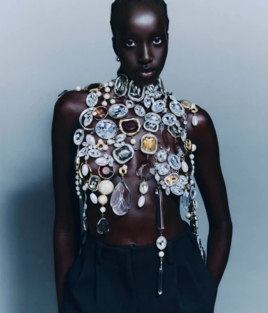 Model Nyaueth Riam wearing elaborate gemstone body jewelry and dark trousers against a plain grey studio backdrop