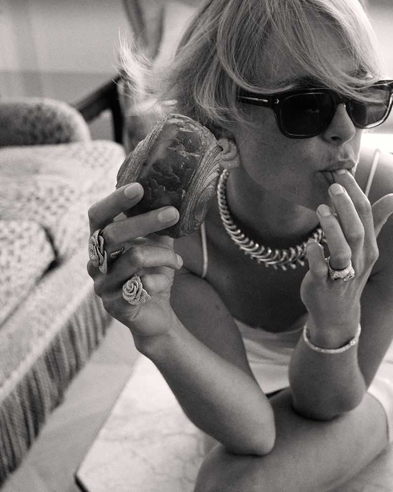 Black and white portrait of a woman with sunglasses, necklace, and rings eating a croissant while sitting on a patterned chair.