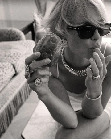 Black and white portrait of a woman with sunglasses, necklace, and rings eating a croissant while sitting on a patterned chair.