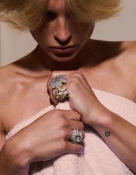 Model with short blonde hair wearing elaborate diamond floral rings and clutching a pale pink textured towel.