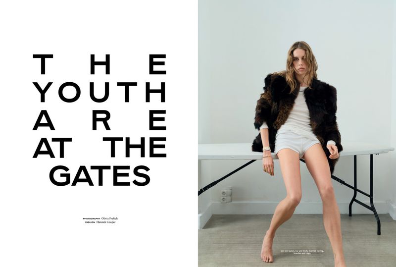 Fashion editorial shot of a woman posing in a fur jacket and shorts in front of a white wall and table with large black text to the left.