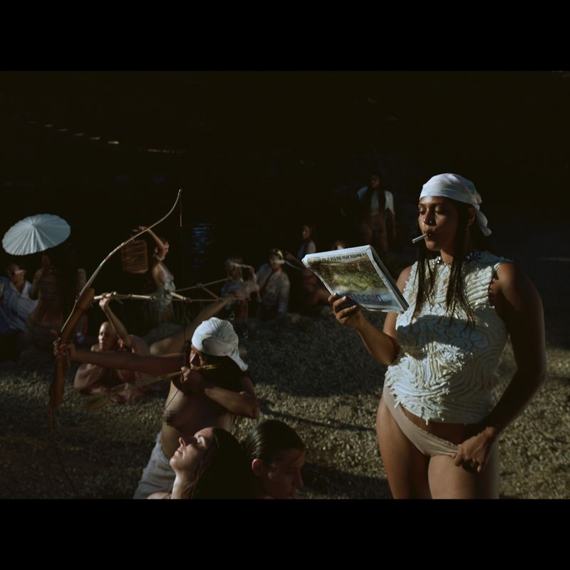 Group of diverse people gathered outdoors, woman in a headscarf reads a magazine, while others are relaxing and engaging in archery nearby.