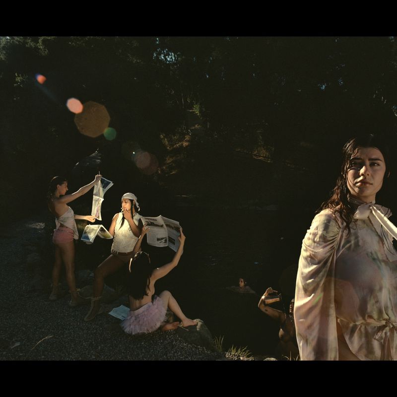 Group of women in outdoor setting, some holding newspapers, one standing in water, wearing gown. Dark environment with natural features.